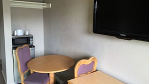 Una habitación pequeña con una mesa redonda de madera, dos sillas moradas acolchadas, un mueble de madera, un televisor montado en la pared y un microondas a la izquierda.