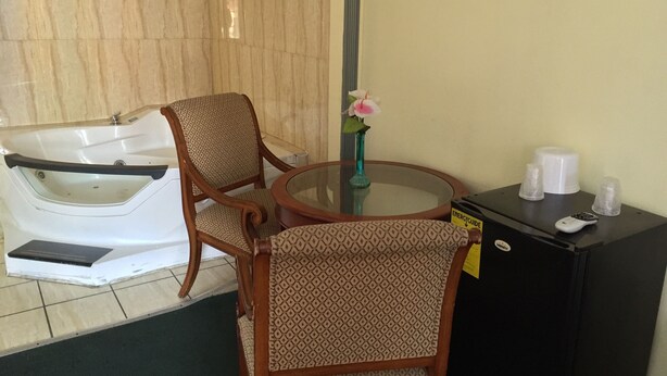 Una habitación con una bañera, dos sillas estampadas, una mesita con un florero y un control remoto, y un mueble negro con una taza blanca y un control remoto.