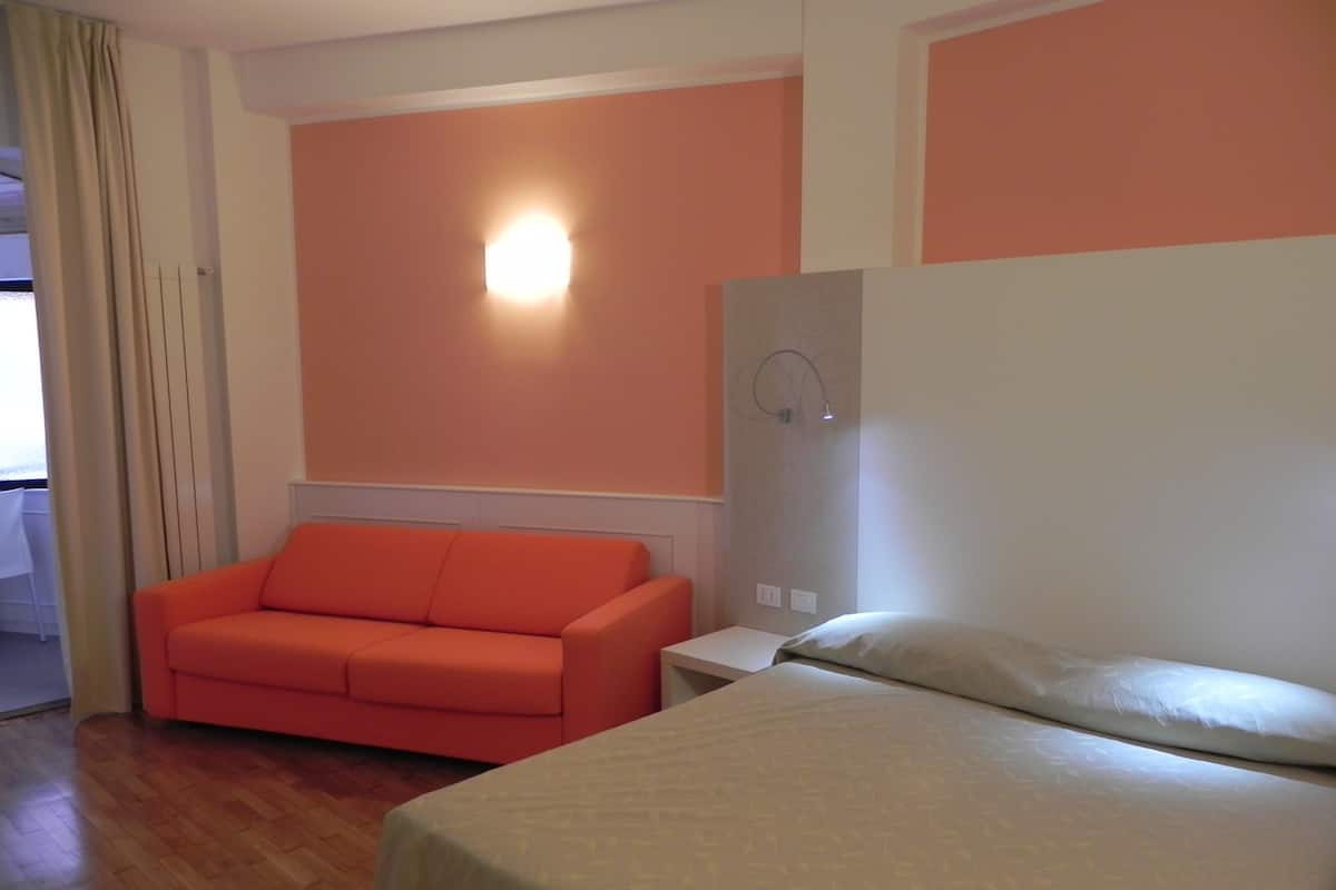 Une chambre d’hôtel avec un lit, un canapé orange, une table de chevet et une lampe fixée au mur.