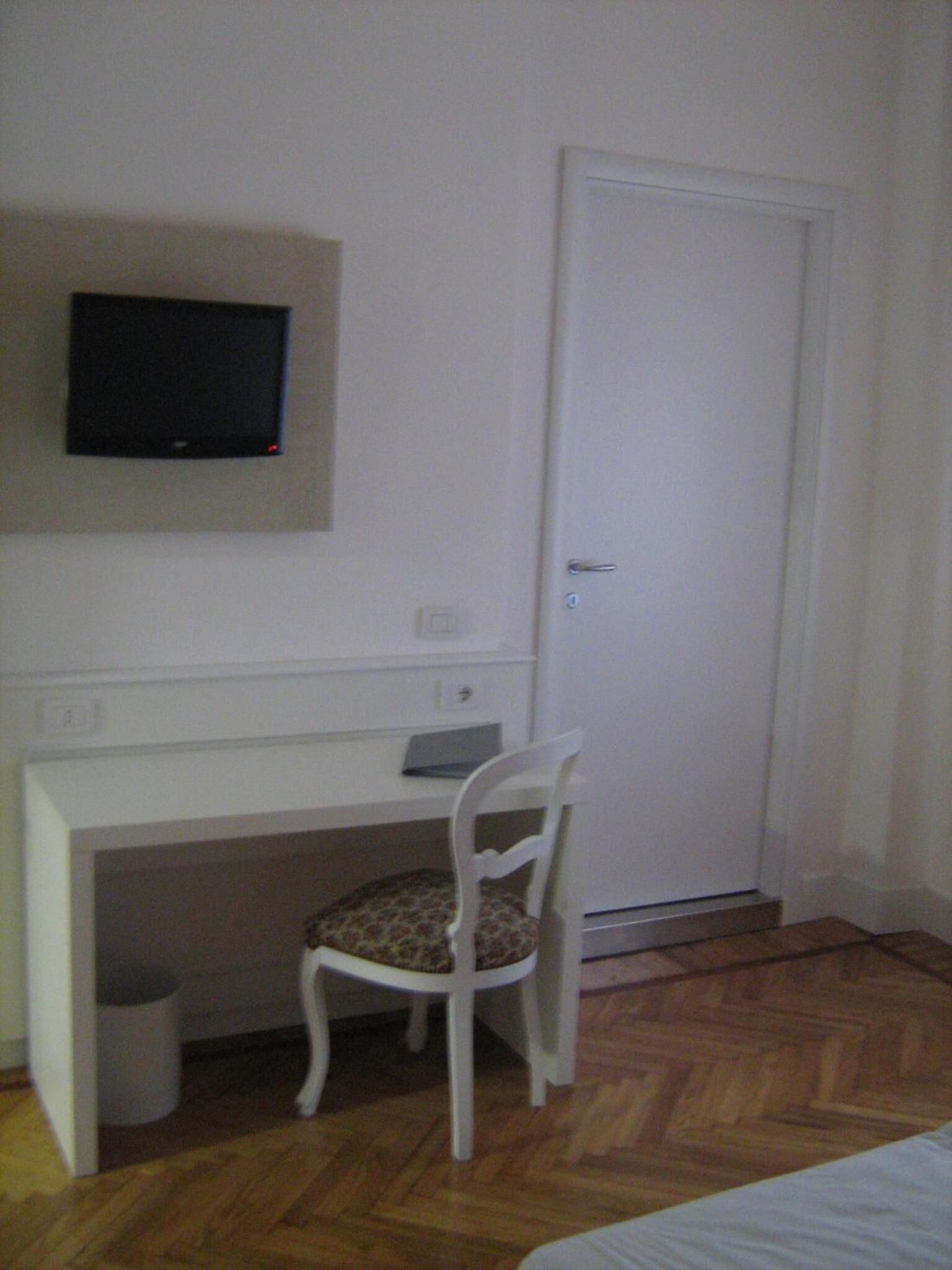 Ein kleiner, sauberer Raum mit einem weißen Schreibtisch, einem Stuhl, einem an der Wand befestigten Fernseher und einer Tür.