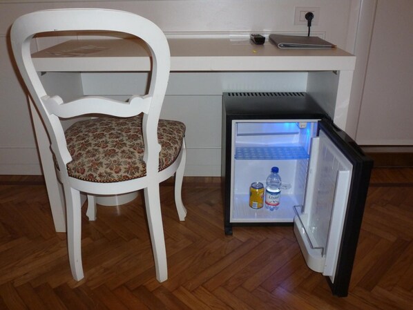 Un refrigerador compacto con una botella de agua y una lata en su interior, situado junto a una silla blanca con un cojín estampado con motivos florales.