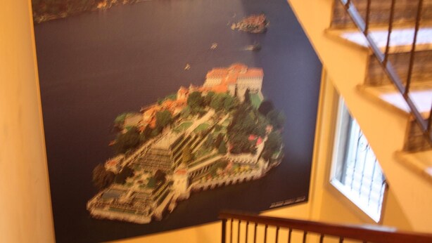 A large, detailed mural of a historical building complex with multiple structures and a surrounding landscape. - Affittacamere Via Mazzini (Stresa)