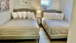 A hotel room with two beds, a small table, and a window with blinds. - Blue Water Inn (Ocean City)