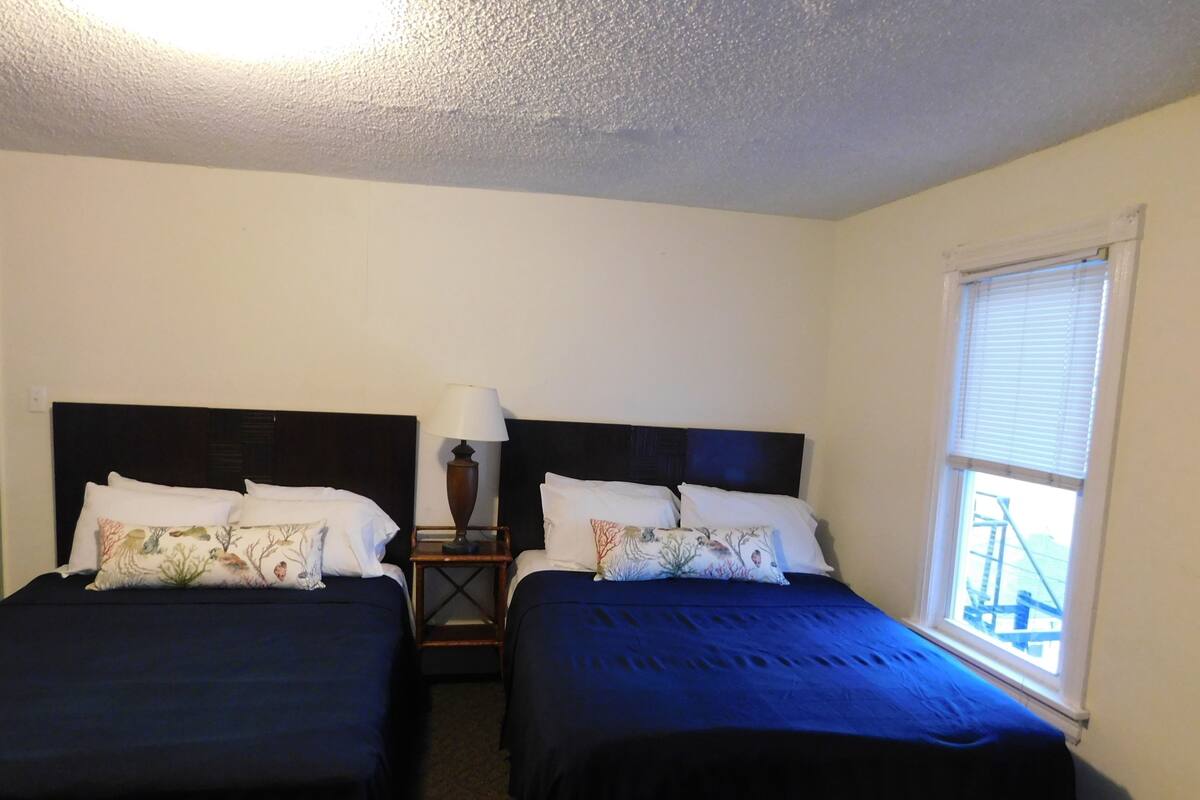 Quarto de hotel com duas camas, uma mesa de cabeceira com abajur e uma janela com persianas.