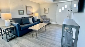 A living room with a sofa, a coffee table, a patterned armchair, and a TV mounted on the wall. - Blue Water Inn (Ocean City)