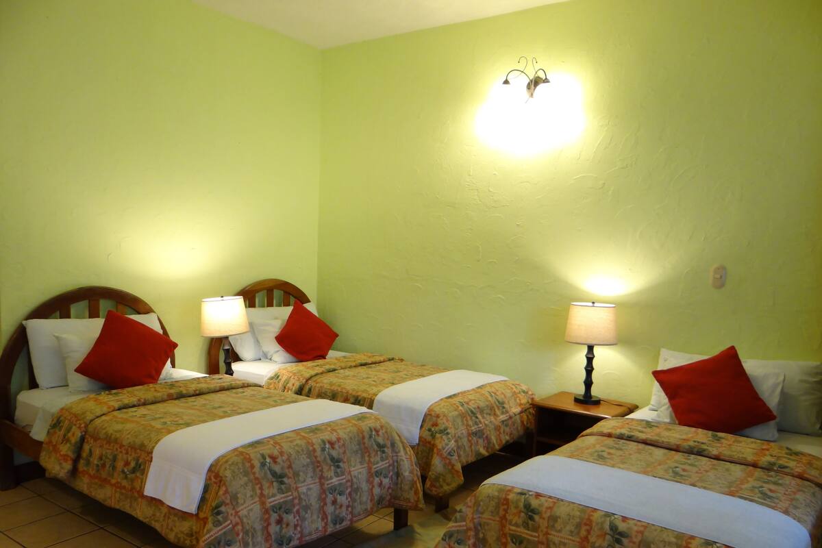 A hotel room with three single beds, each with a floral bedspread and red pillows. There are two bedside lamps and a wall-mounted light fixture.