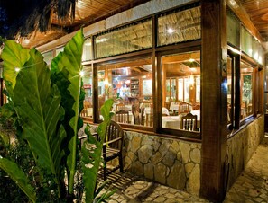 An outdoor restaurant with stone walls, wooden furniture, and large windows. - Hotel Jaguar Inn Tikal (Flores)