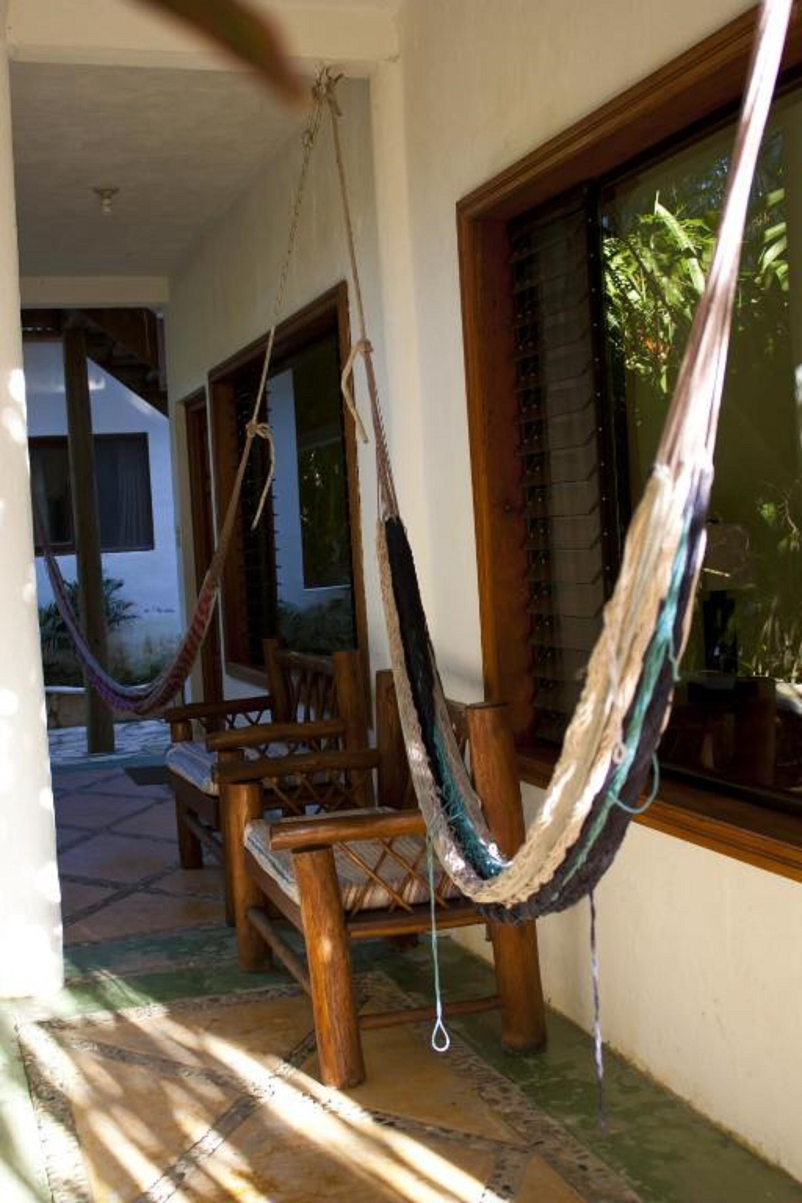 A hammock hanging between wooden chairs in a room with a large window and a view of greenery outside.