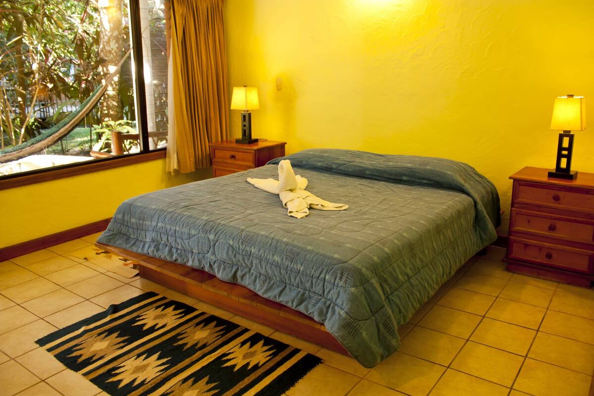 A hotel room with a bed, two bedside tables with lamps, a window with curtains, a view of greenery outside, and a patterned rug on the floor.