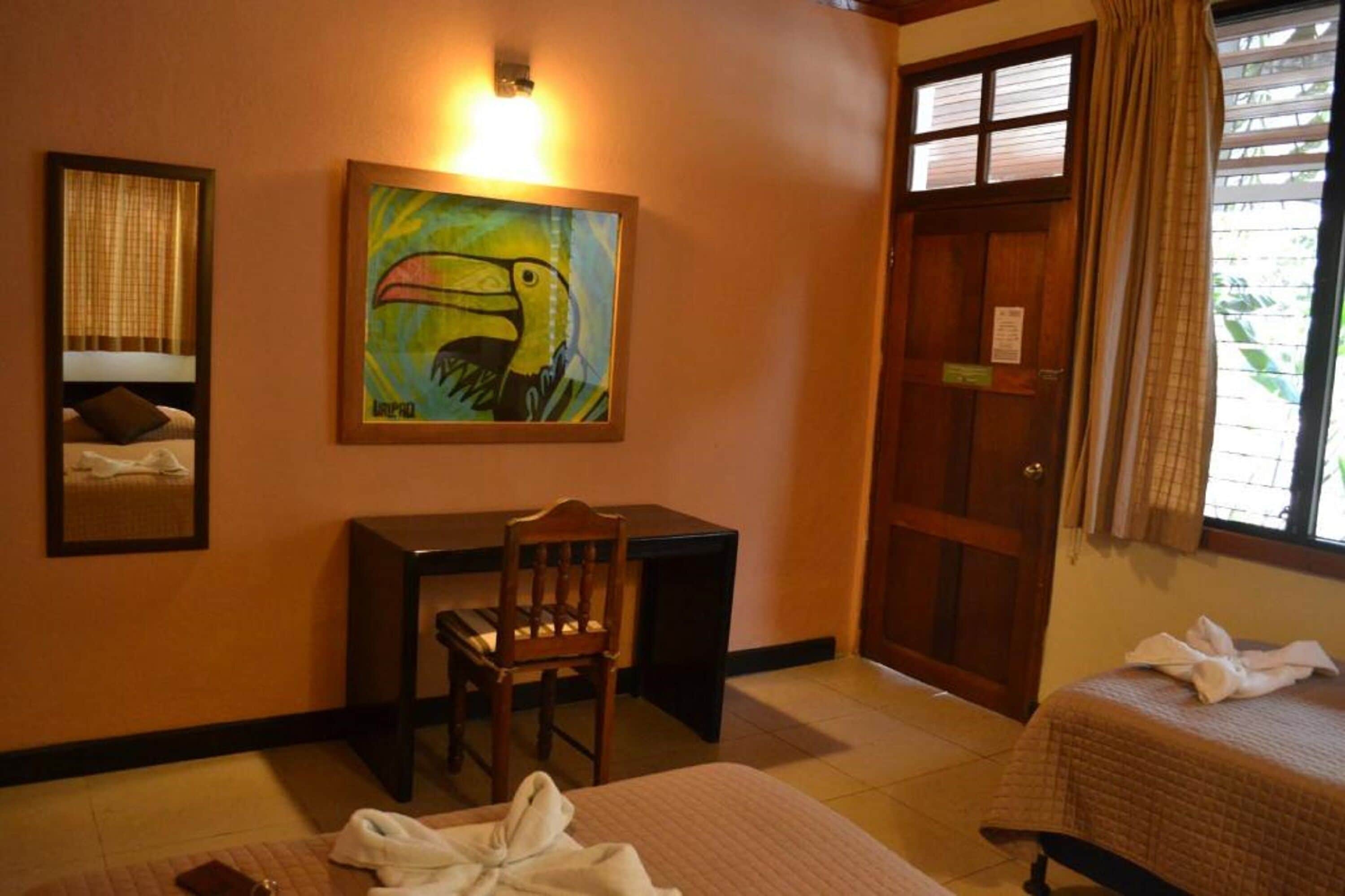 A hotel room with a bed, a desk, a chair, a painting of a toucan, and a door.