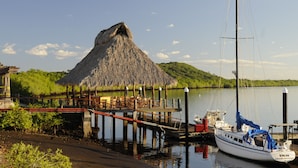 Um cais com uma construção de telhado de palha, um barco e um lago calmo.