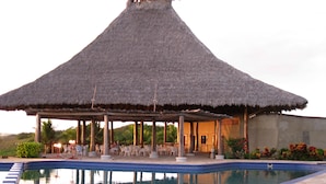 Uma área de piscina com um pavilhão de telhado de palha, piscina de pedra e mobiliário de exterior.