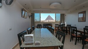 A dining area with a long table, chairs, and a view of pyramids through large windows. - Guardian Guest House (Giza)
