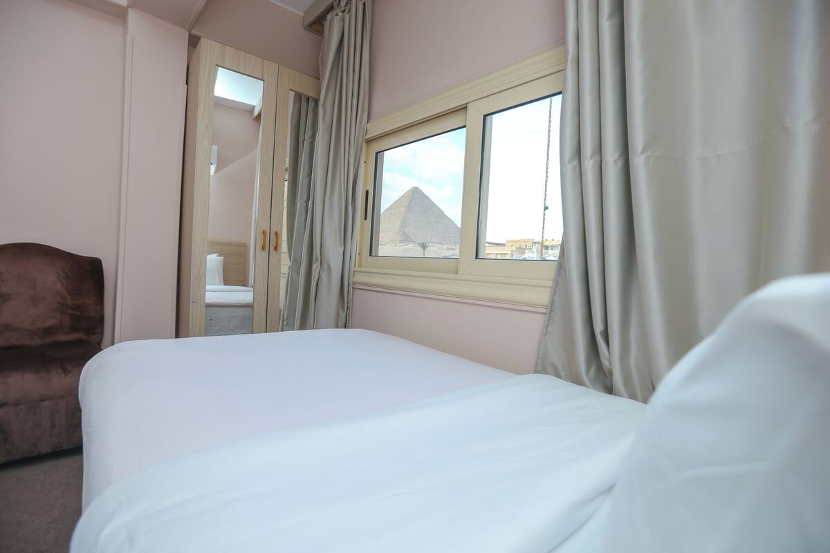 A bedroom with a bed, a chair, a window with a view of a pyramid, and a door leading to another room.