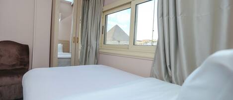A bedroom with a bed, a chair, a window with a view of a pyramid, and a door leading to another room.