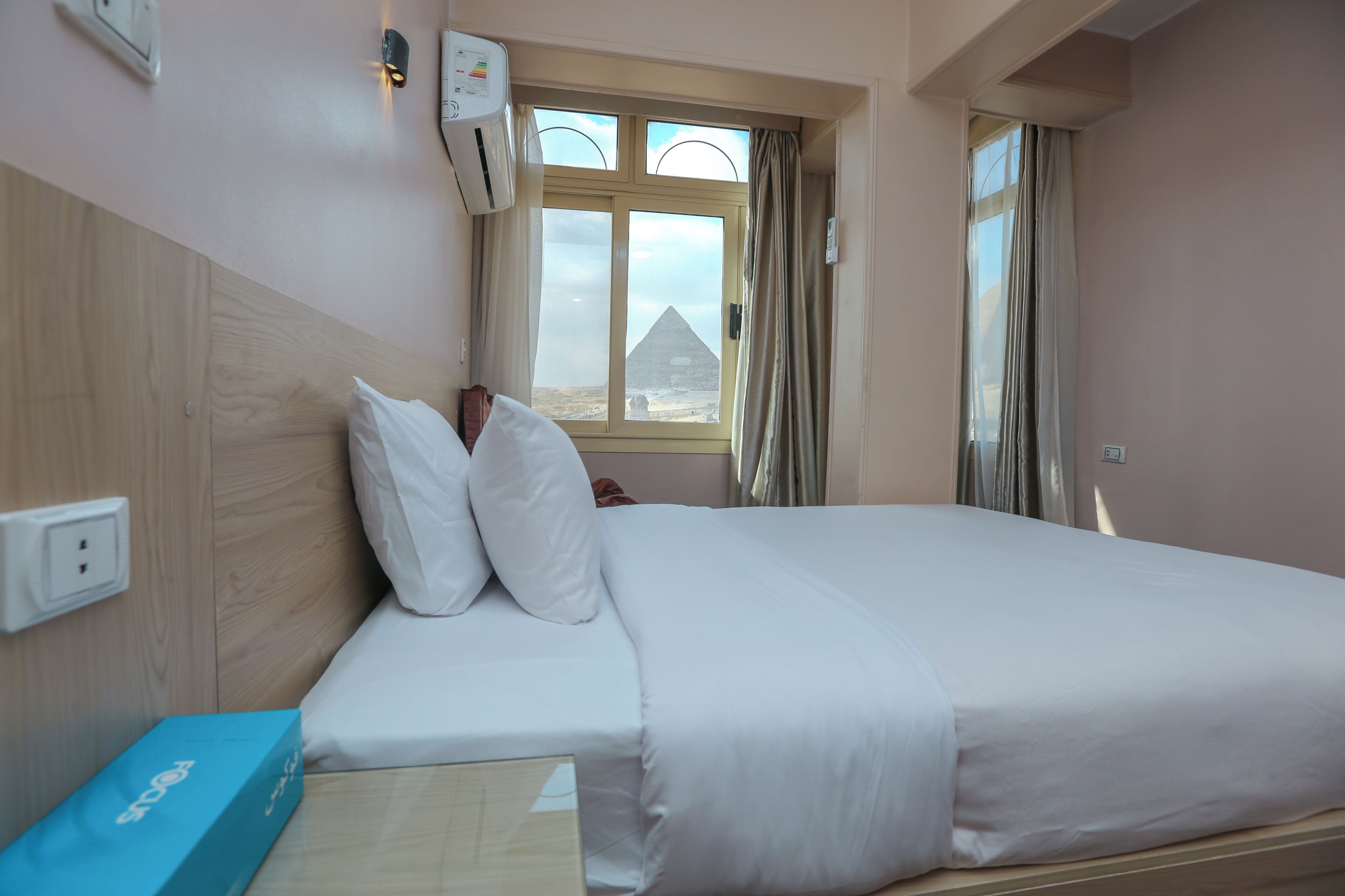 Ein Hotelzimmer mit Bett, einem Fenster mit Vorhängen, einer Klimaanlage und einer blauen Schachtel auf dem Nachttisch.