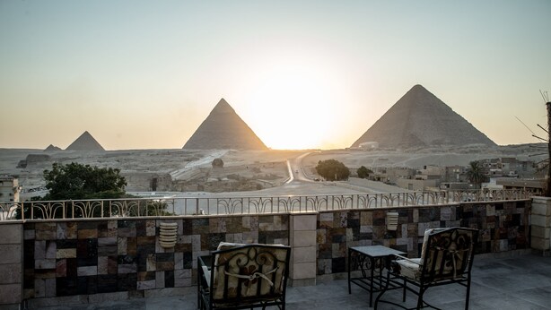 A terrace with two chairs and a table, overlooking a desert landscape with pyramids and a road leading towards the pyramids. - Guardian Guest House (Giza)