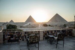 Una terraza con dos sillas y una mesa, con vistas a un paisaje desértico que incluye pirámides y un camino que lleva hacia ellas.