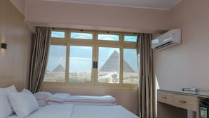 A hotel room with a bed, a desk, an air conditioning unit, and a view of the pyramids. - Guardian Guest House (Giza)