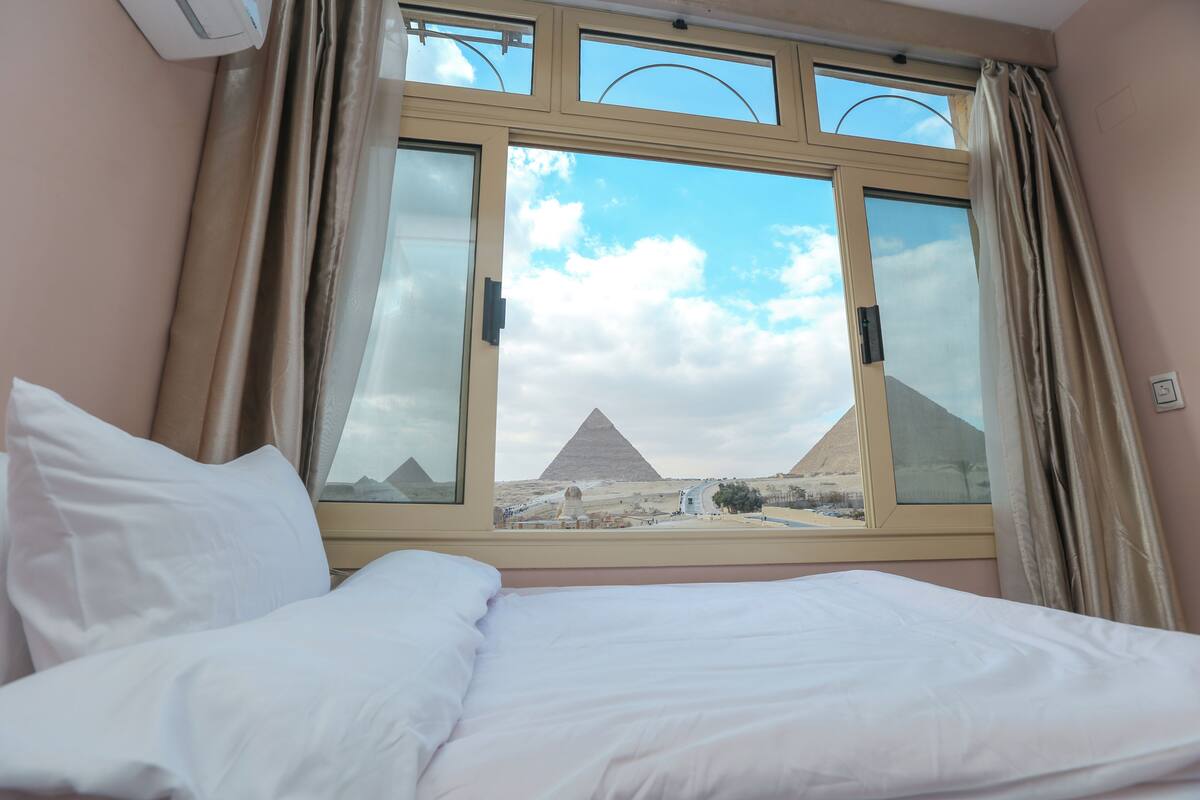 A hotel room with a bed, a window with curtains, and a view of the pyramids.