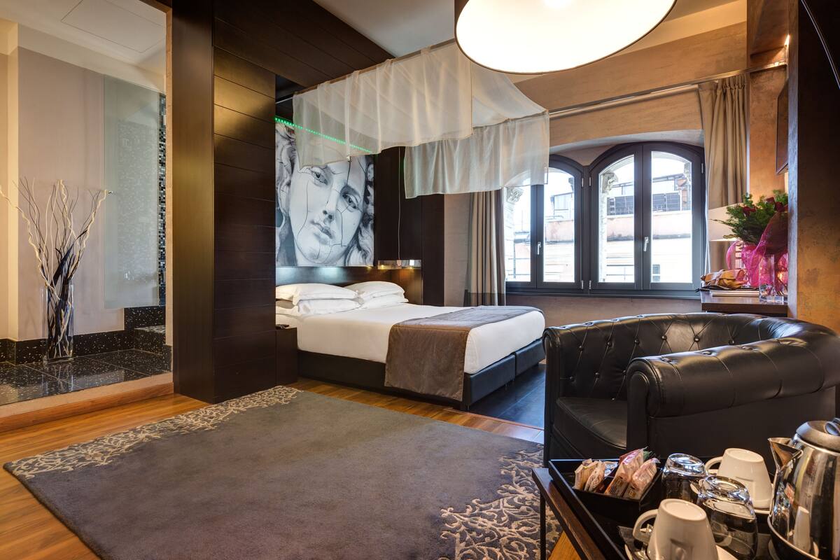 A modern hotel room with a large bed, a leather sofa, a dining table with breakfast items, and a large window with a view of buildings.