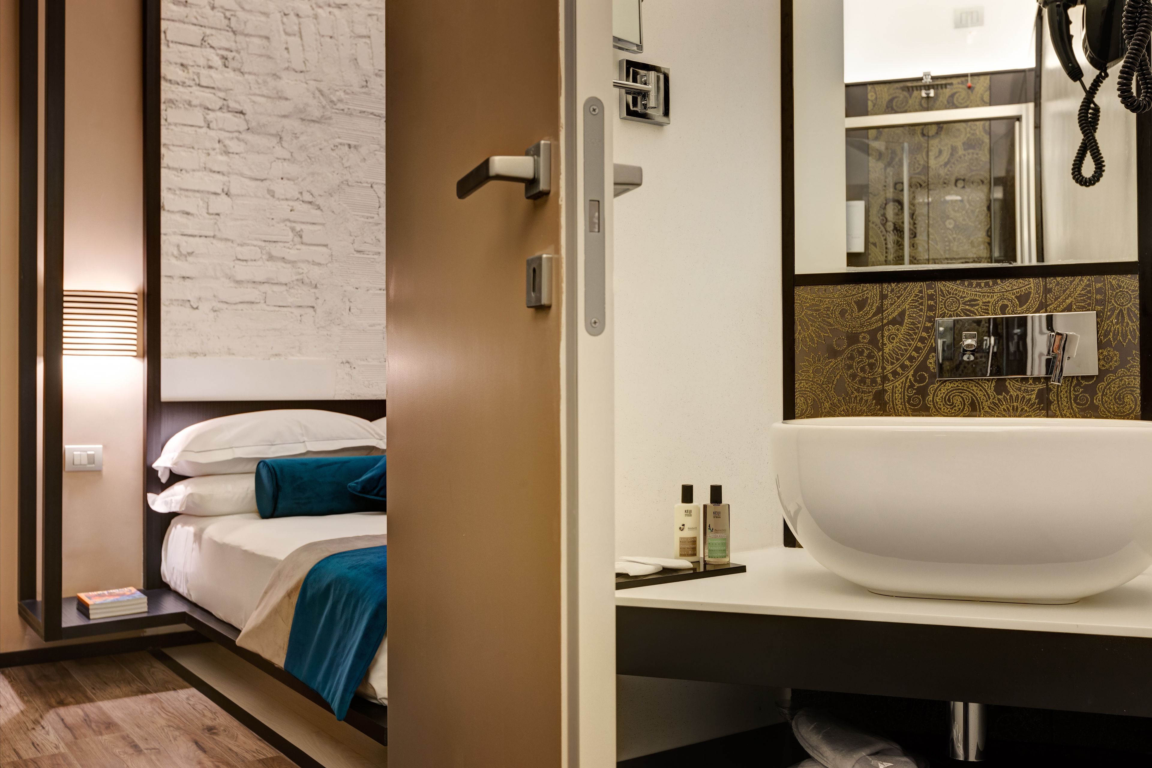 A hotel room with a bed, a bedside table with a book, a large mirror, and a bathroom with a white sink and a large mirror.