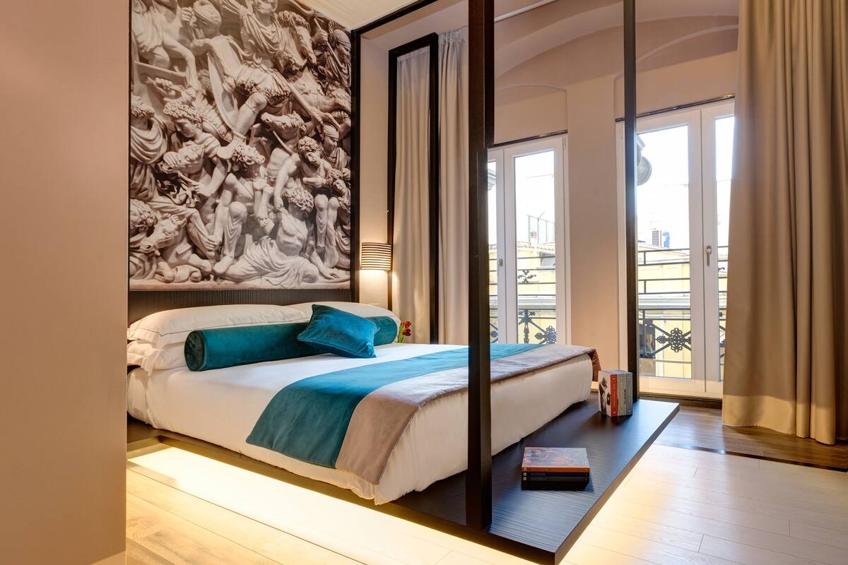 A modern hotel room with a large bed, a view of the city, and a unique wall sculpture.