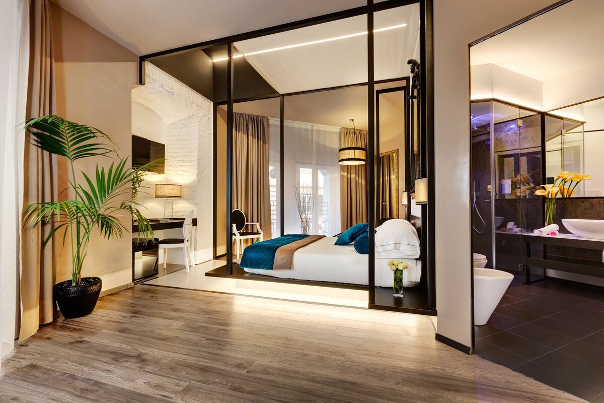 A modern hotel room with a large bed, a sitting area, a bathroom with a mirror, and a potted plant.