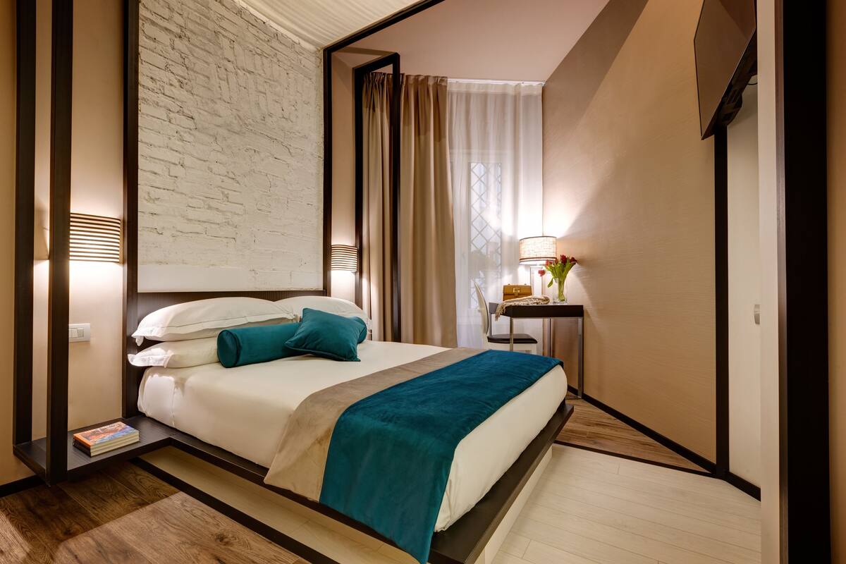 A hotel room with a bed, a bedside table with a lamp, a small table with a vase of flowers, and a brick wall.