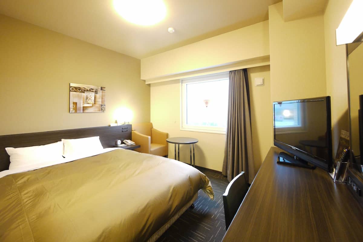 A hotel room with a large bed, a desk with a flat-screen TV, a small round table, and a window with curtains.