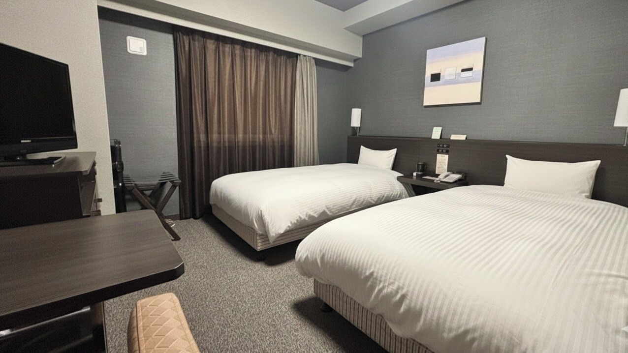 A hotel room with two beds, a desk, a television, and a view of the cityscape.