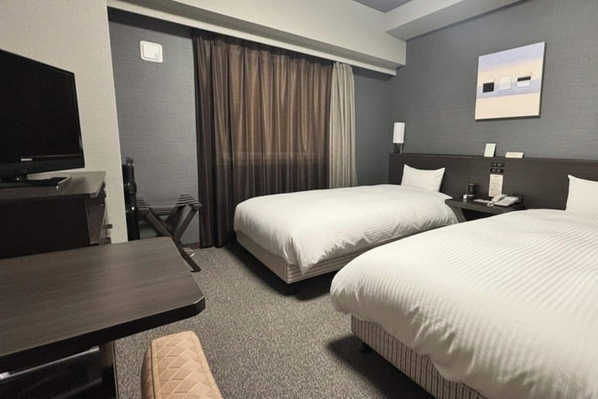 A hotel room with two beds, a desk, a television, and a view of the cityscape.