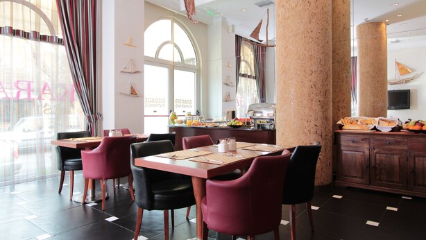 Un ristorante con angolo colazione, sedie rosse e nere, e un buffet con frutta e pasticceria.