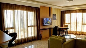 A hotel room with a seating area, a television mounted on the wall, and large windows with curtains.