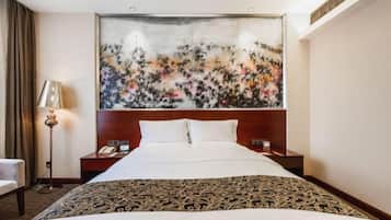 A hotel room with a large bed, a headboard with artwork, a bedside table with a lamp, and a chair.