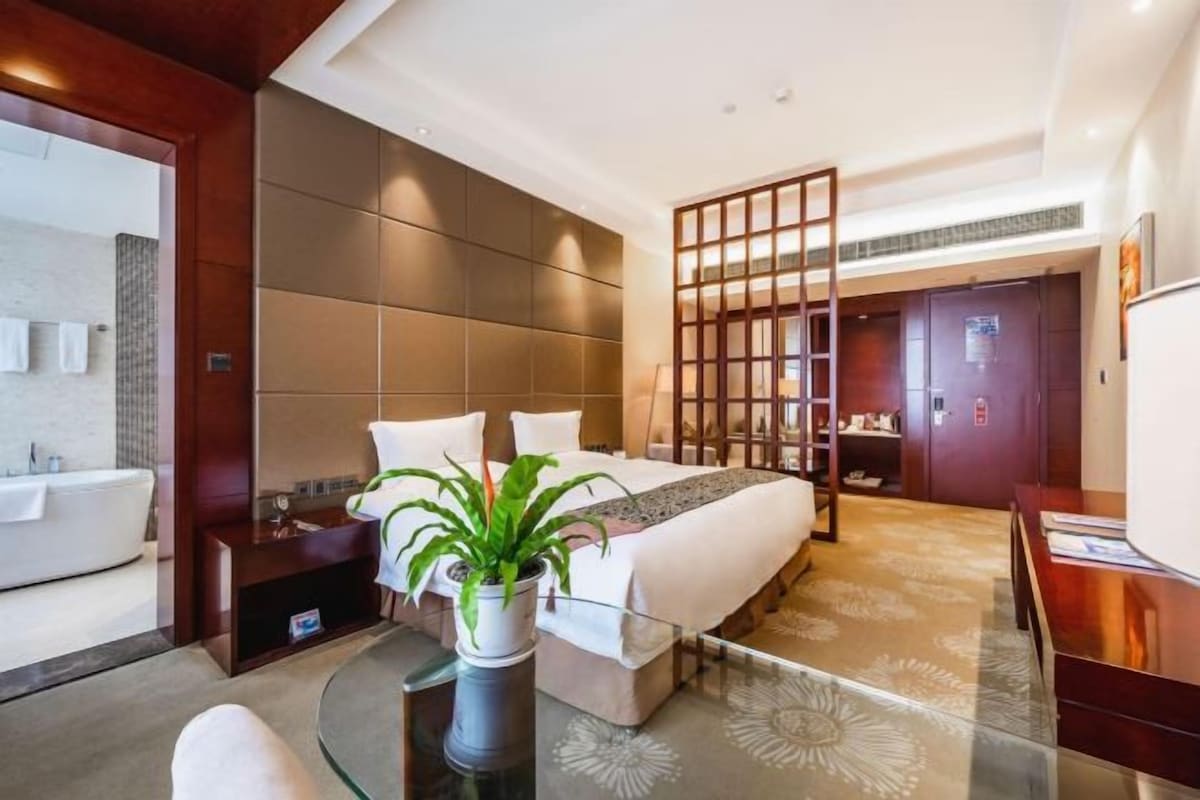 Ein modernes Hotelzimmer mit einem großen Bett, einem Schreibtisch, einem Badezimmer mit Badewanne und einer Zimmerpflanze.