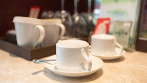 Two white cups on saucers on a table, with a tray of similar cups and a glass in the background. - Intercity Hotel (Baoji)