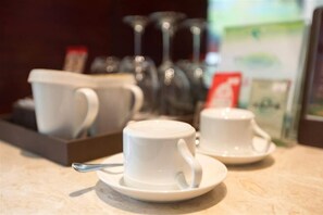 Dos tazas blancas sobre platillos en una mesa, con una bandeja de tazas similares y un vaso al fondo.