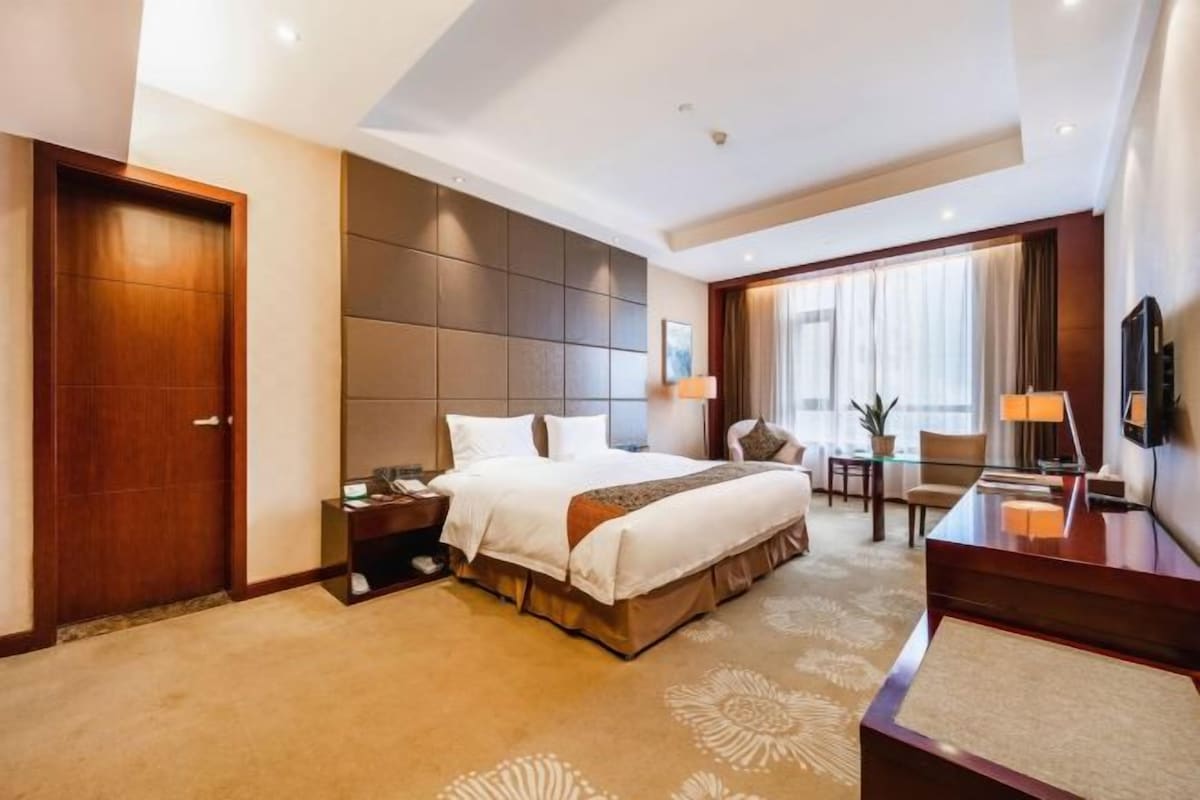 Ein Hotelzimmer mit einem großen Bett, einem Schreibtisch, einem Sessel, einem Fernseher und einem Fenster mit Vorhängen.