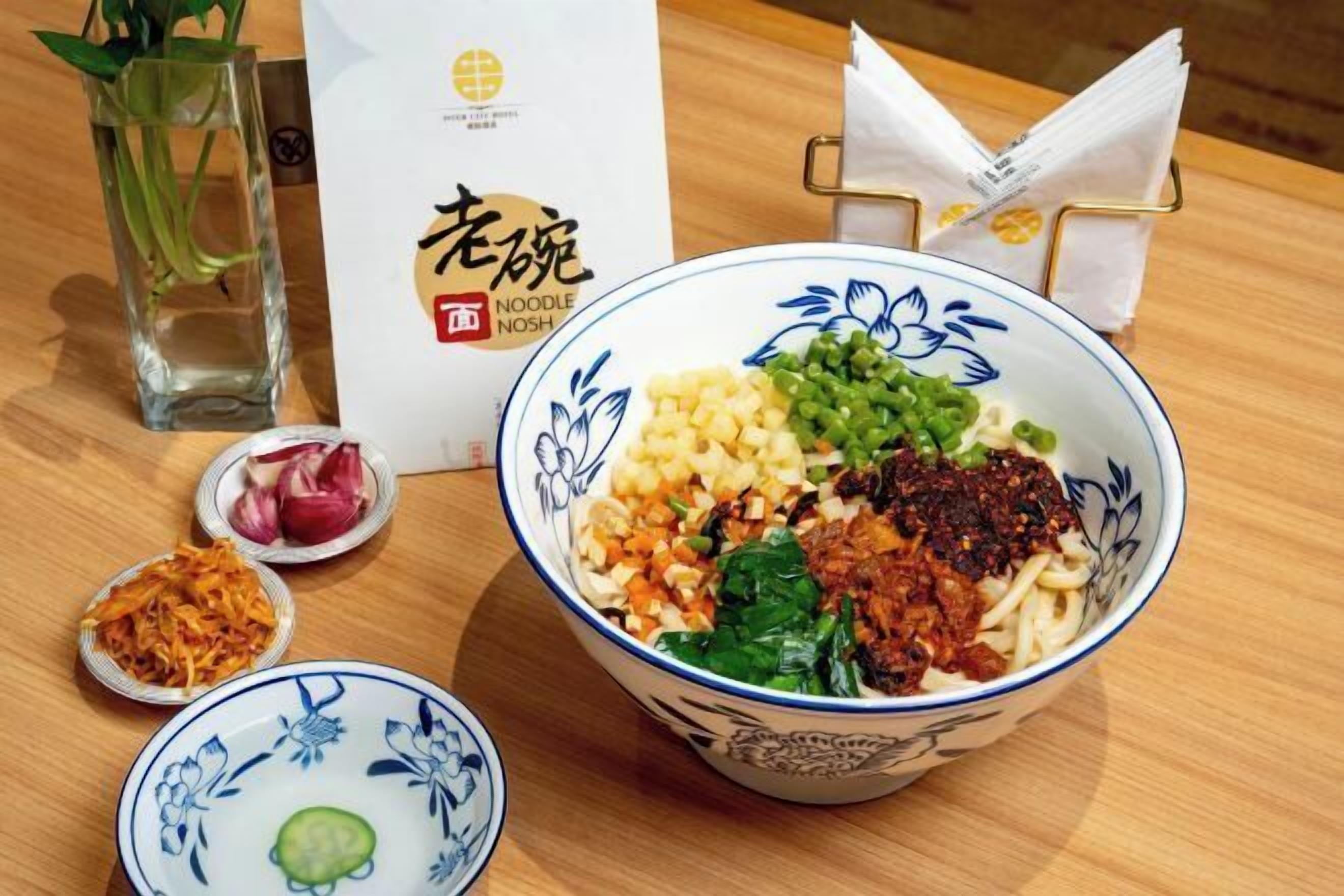 A bowl of noodle soup with various toppings, a menu, and a small dish with a cucumber slice.