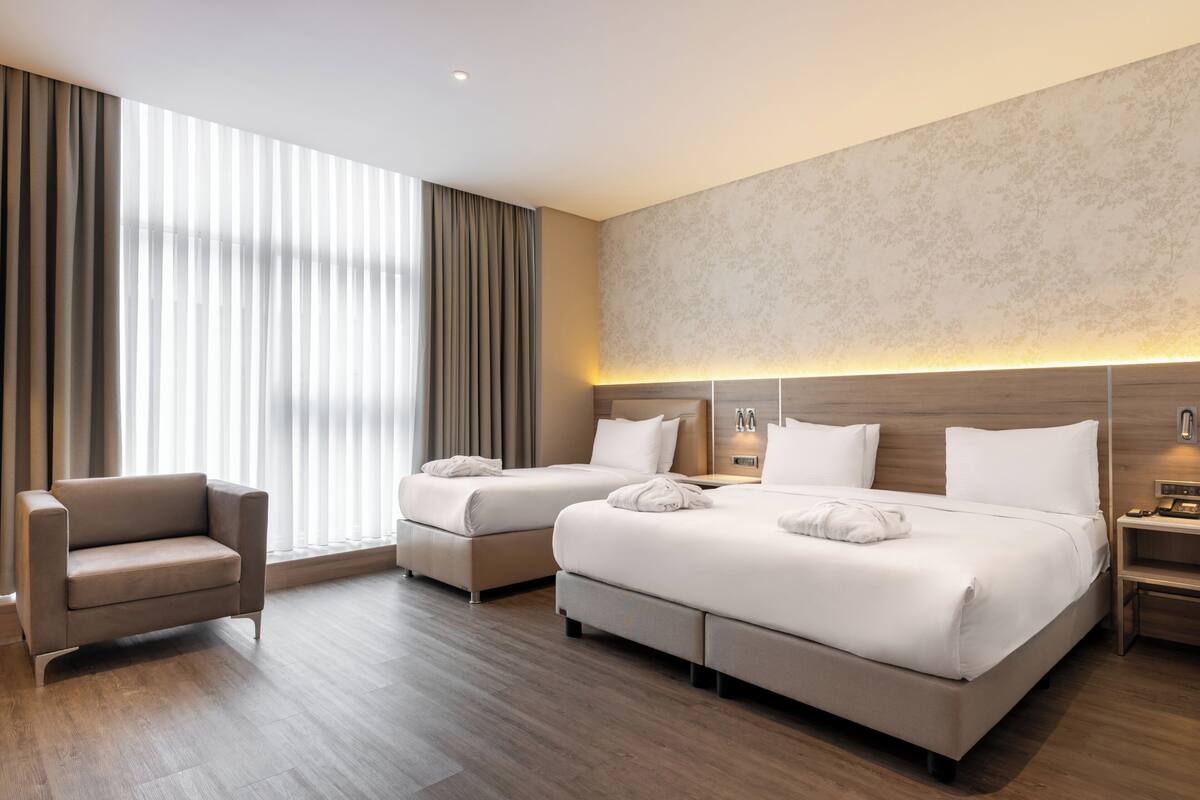 Habitación de hotel con dos camas, un sofá, una mesita de noche y un ventanal con persianas.