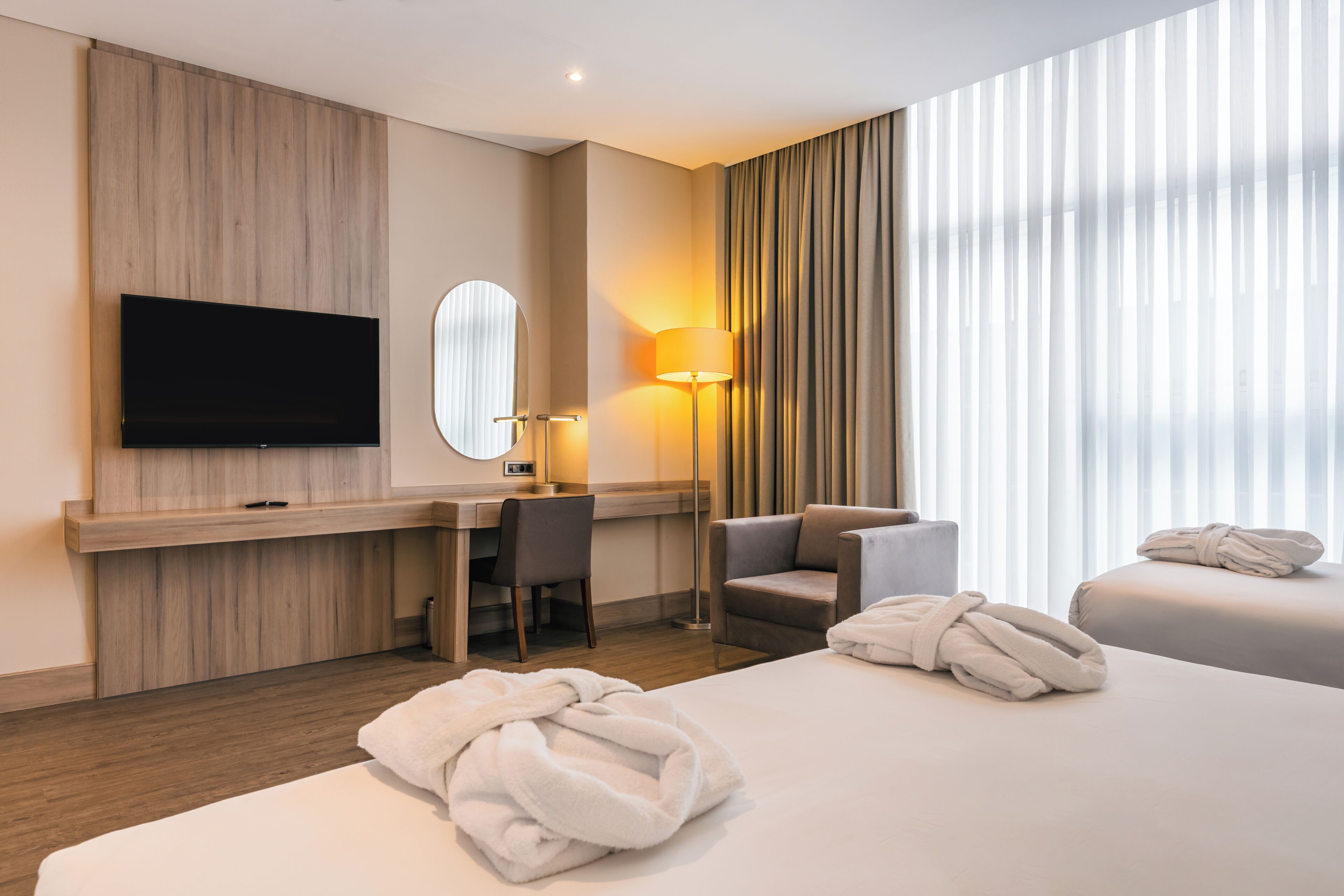 A hotel room with a flat-screen TV, a desk with a chair, a sofa, two beds, and a large window with curtains.