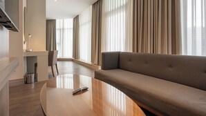 A modern hotel room with a curved wooden coffee table, a sofa, a dining area with a chair, and large windows with curtains. - Ramada Encore by Wyndham Istanbul Bayrampasa (Istanbul)