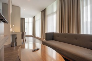 Ein modernes Hotelzimmer mit einem gewölbten Holztisch, einer Couch, einem Essbereich mit einem Stuhl und großen Fenstern mit Vorhängen.