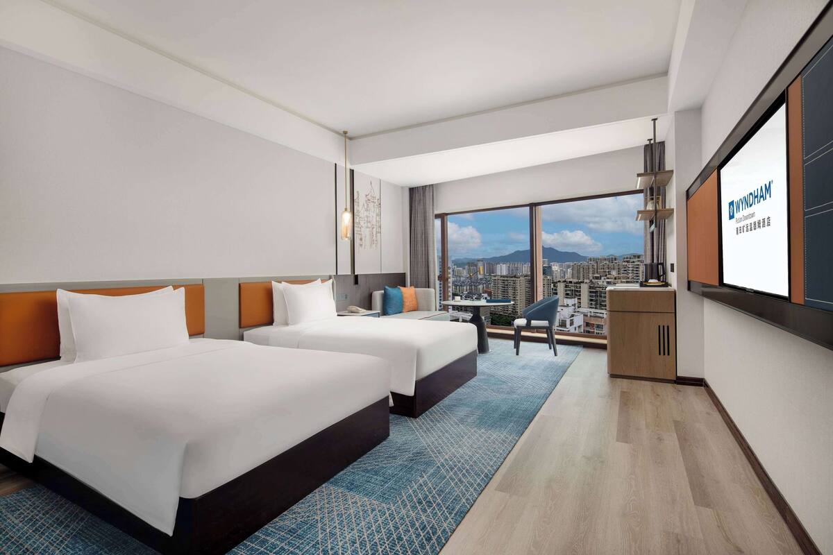 Habitación de hotel con dos camas, un escritorio y un ventanal grande con vistas a la ciudad.