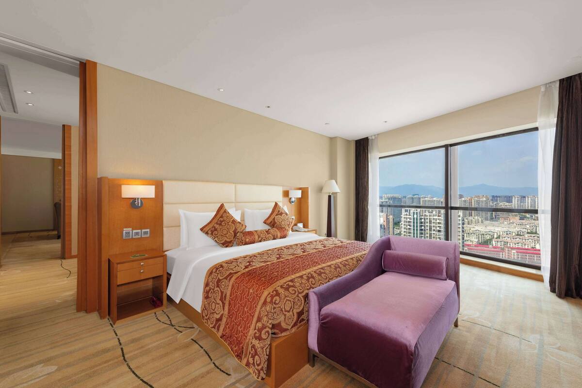 Habitación de hotel con una cama grande, una mesita de noche, un sillón púrpura y vistas a la ciudad a través de amplios ventanales.
