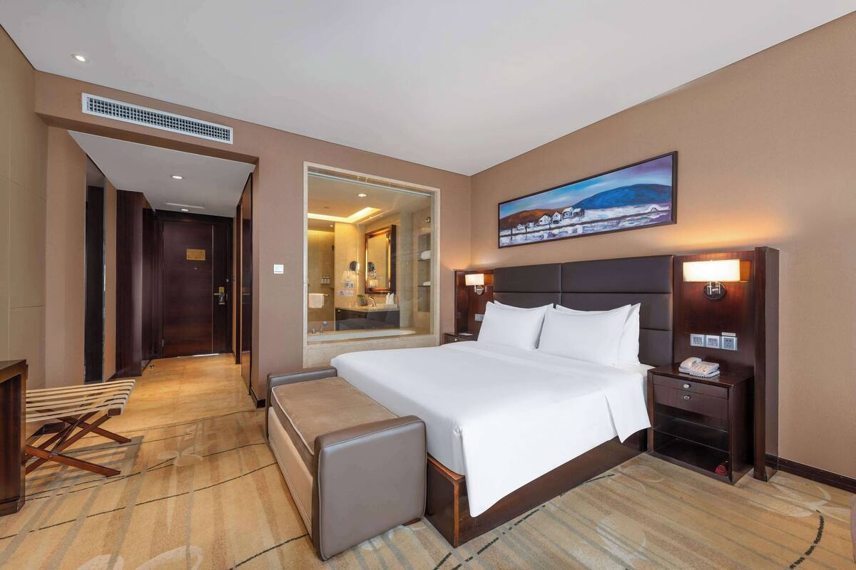 Habitación de hotel con una cama grande, una mesita de noche con lámpara, un pequeño rincón de descanso y vistas al baño a través de una puerta de cristal.
