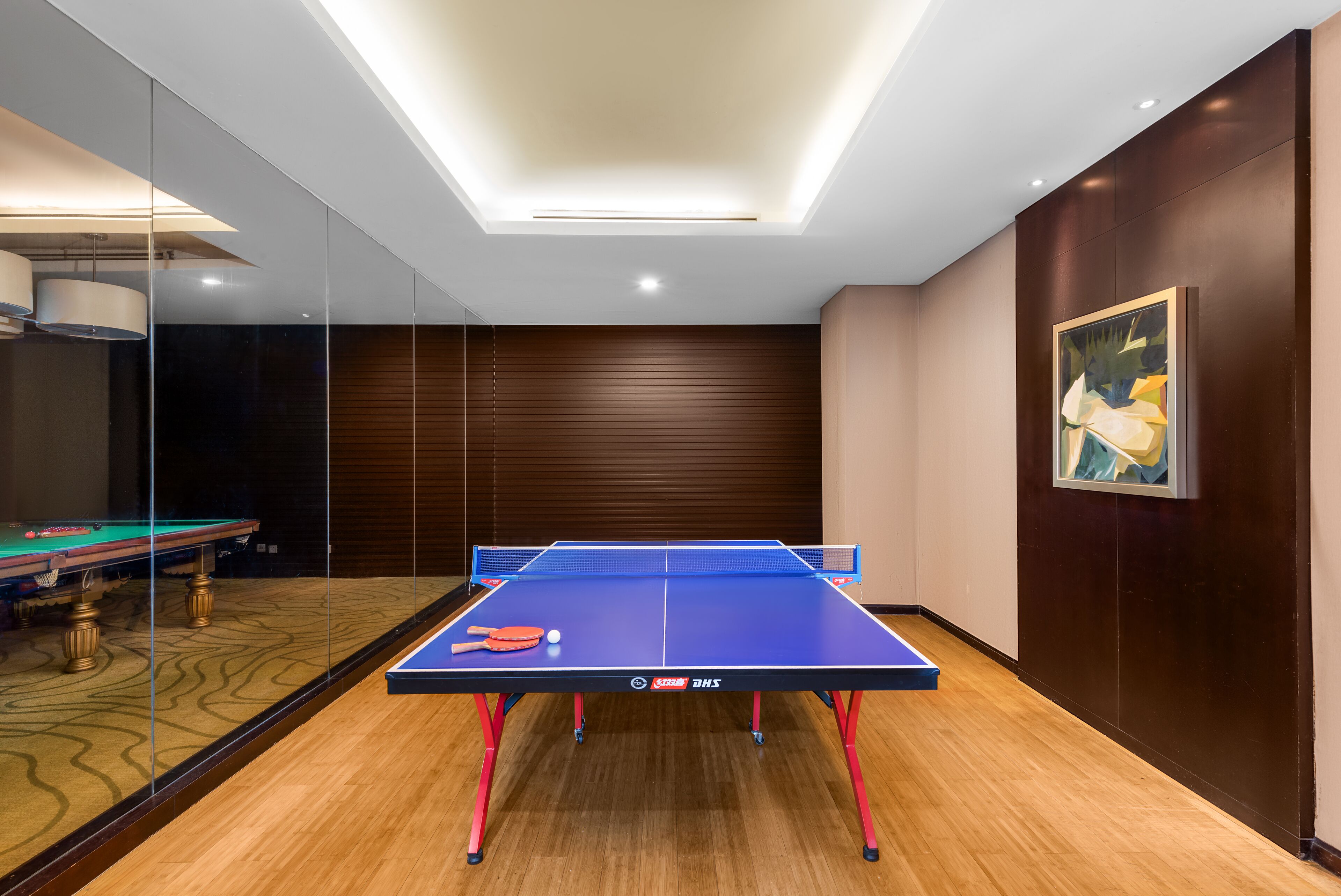 A ping pong table in a modern room with wooden flooring, a glass partition, and a ceiling light fixture.