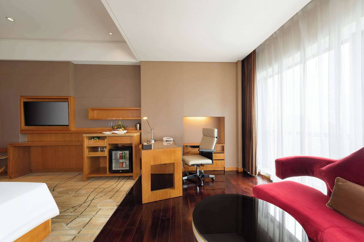 Habitación de hotel con cama, escritorio con computadora, televisor, sofá rojo y ventana grande con cortinas.