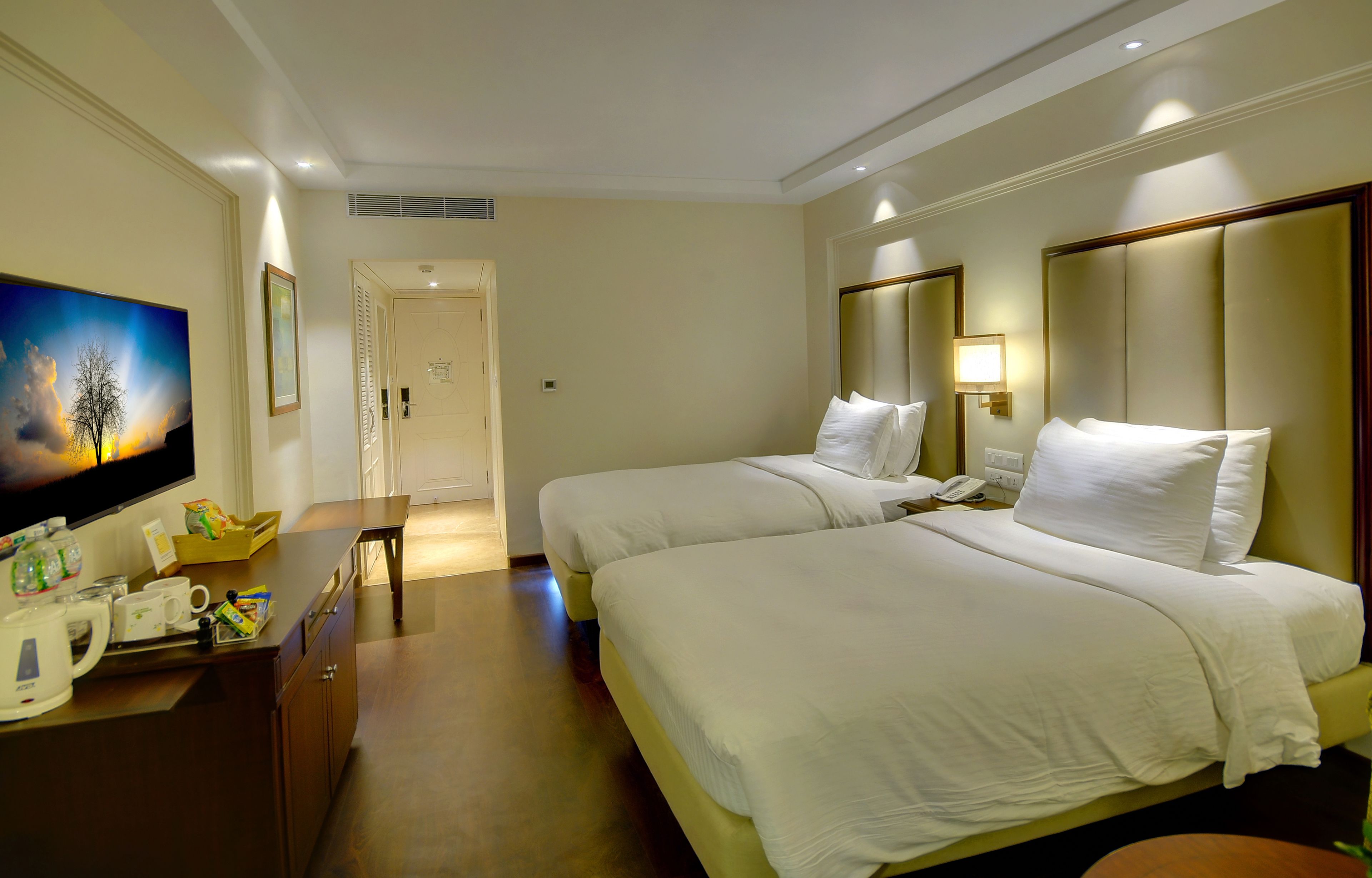 Une chambre d’hôtel avec deux lits, une télévision, un bureau équipé d’une bouilloire et d’une machine à café, et une porte donnant sur une salle de bain.
